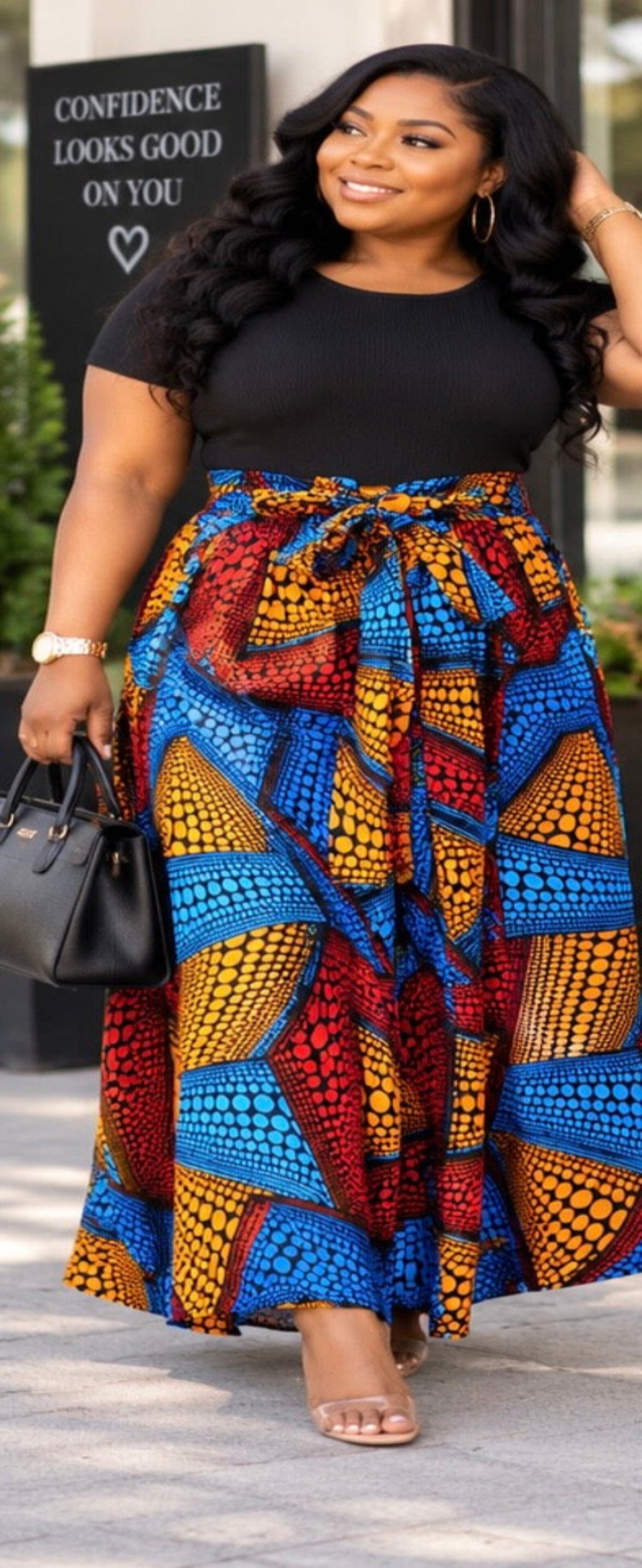 A woman wearing a black short-sleeve top and a high-waisted, tie-waist maxi skirt with a bold multicolor African print in blue, red, yellow, and orange. She is carrying a black handbag and wearing clear high-heeled sandals, standing outdoors in front of a building with a sign.