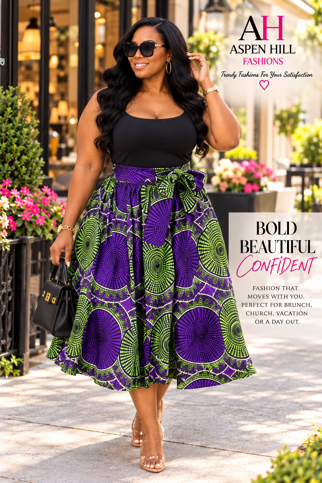 A woman models a sleeveless black top and a high-waisted midi skirt featuring a bold purple and green circular African wax print. She is walking outdoors on a paved street with storefronts and flowers in the background.