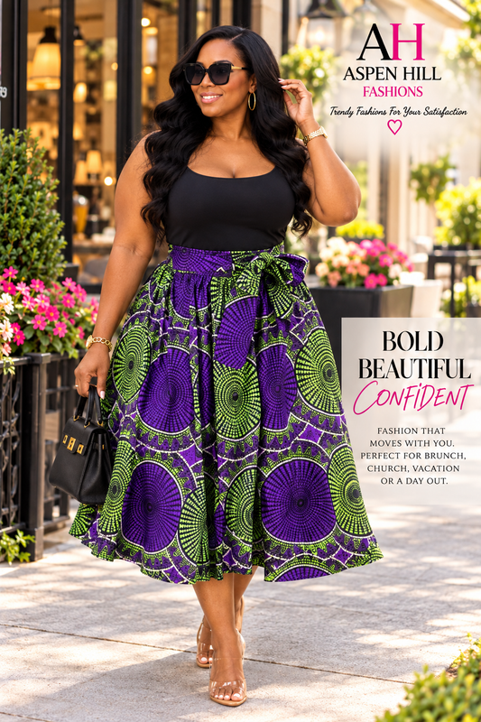 A woman models a sleeveless black top and a high-waisted midi skirt featuring a bold purple and green circular African wax print. She is walking outdoors on a paved street with storefronts and flowers in the background.