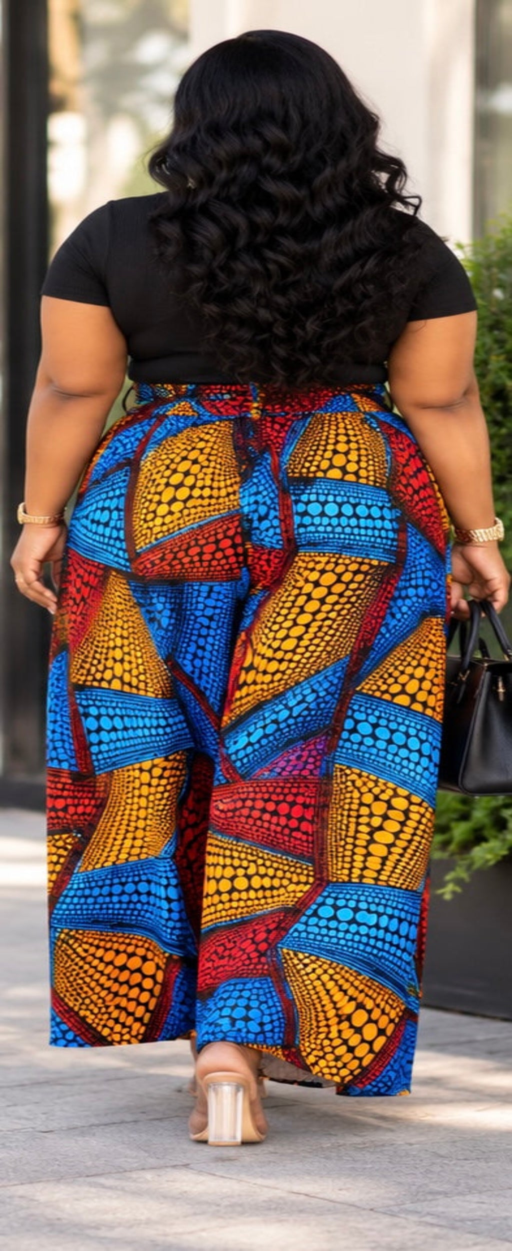 A woman viewed from behind wearing a black short-sleeve top and a very wide-leg, high-waisted skirt with a bold multicolor African wax print pattern in blue, red, yellow, and orange, walking outdoors.
